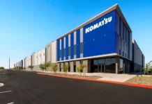 Komatsu Strengthens U.S. Network with Arizona Parts Center Exterior of a modern Komatsu office building with blue vertical panels, glass doors, and a paved parking lot under a clear blue sky.