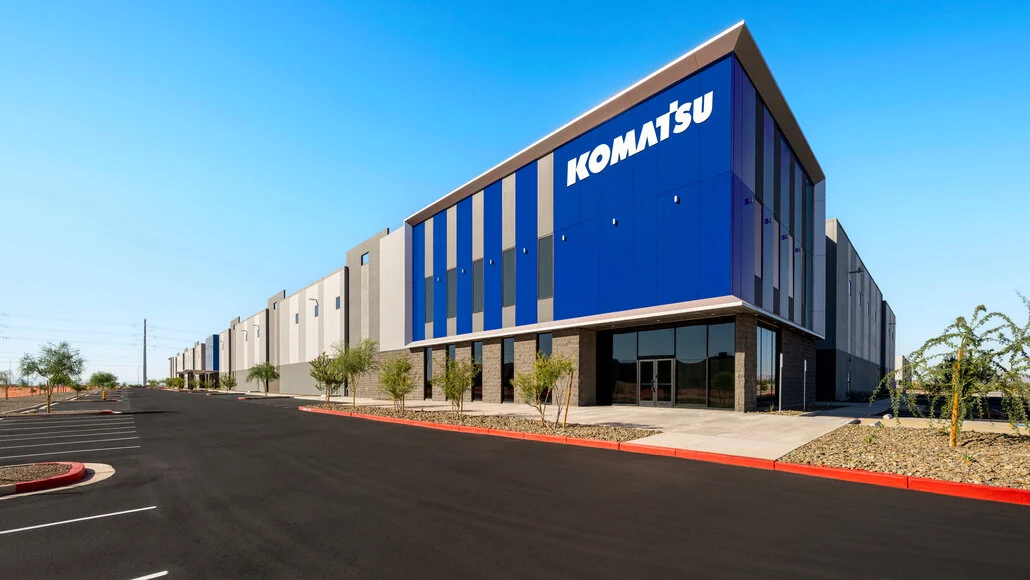 Exterior of a modern Komatsu office building with blue vertical panels, glass doors, and a paved parking lot under a clear blue sky.