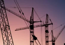 Why Equipment Down Times Become Costly in Road Construction Projects Silhouettes of tall construction cranes against a pink and purple sunset sky, with cables crisscrossing overhead.