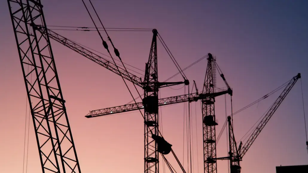 Silhouettes of tall construction cranes against a pink and purple sunset sky, with cables crisscrossing overhead.