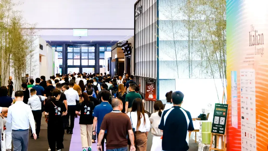 Busy indoor exhibition hall with a dense crowd walking down a corridor between booths and colorful walls on the right.